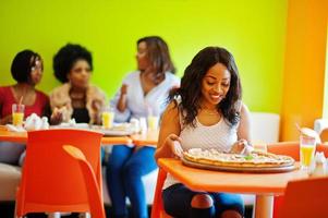 African woman with pizza sitting at restaurant against dark skinned girls. photo