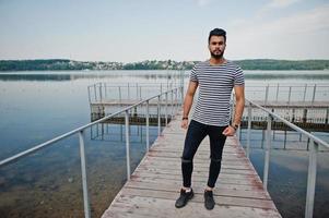 apuesto modelo de hombre de barba árabe alto en camisa desnuda posada al aire libre en el muelle del lago. chico árabe de moda. foto