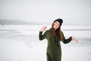 Funny girl wear on long green sweatshirt, jeans and black headwear, at frozen lake in winter day. photo