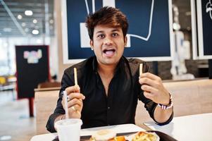 Funny and stylish indian man sitting at fast food cafe and eating french fries, show his tongue. photo