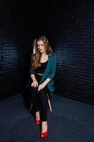 hermosa chica morena vestida con una chaqueta negra y verde con tacones rojos, sentada y posando en una silla en el estudio contra una pared de ladrillo oscuro. retrato de modelo de estudio. foto