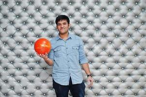 Stylish asian man in jeans shirt standing with bowling ball at hand against silver wall background. photo