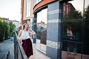 Fashionable and beautiful blonde model girl in stylish red velvet velour skirt, white blouse, posed with phone and earphones against windows of city building. photo