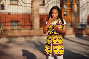 elegantes mujeres afroamericanas con chaqueta amarilla posaron en la calle en un día soleado con el teléfono móvil a mano. cara de asombro foto