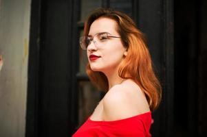 Attractive redhaired woman in eyeglasses, wear on red blouse posing at street against old wooden door. photo