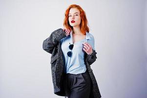 Portrait of a fantastic redheaded girl in blue shirt, grey overcoat posing with sunglasses in the studio. photo