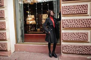 Portrait of a curly haired african woman wearing fashionable black coat and red turtleneck posing outdoor. photo