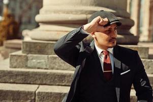 Portrait of retro 1920s english arabian business man wearing dark suit, tie and flat cap near old columns. photo