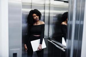 Fashionable young beautiful african american business woman with afro hairstyle wear in elegant black, stand at elevator with phone and laptop at hands. photo