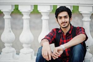 Young indian student man at checkered shirt and jeans with backpack posed on evening city against column. photo