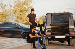 Two asian brothers man wear on all black posed near suv cars and smoke hookah. photo