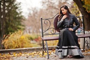 linda chica india con vestido de sari negro y chaqueta de cuero posada al aire libre en la calle de otoño y sentada en un banco. foto