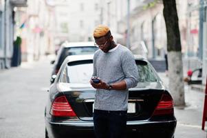 Elegante chico afroamericano con suéter gris y anteojos posó en la calle contra un auto de negocios negro y hablando por teléfono. chico negro de moda. foto