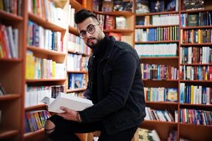 estudiante árabe alto e inteligente, vestido con chaqueta negra de jeans y anteojos, en la biblioteca con un libro en las manos. foto