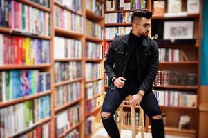 Tall smart arab student man, wear on black jeans jacket and eyeglasses, at library sitting on chair against book shelves. photo