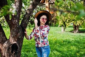 Spring portrait of brunette girl in pink glasses and hat at green blossom garden. photo