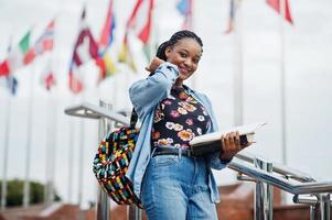 African student female posed with backpack and school items on yard of university, against flags of different countries. photo