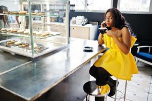 Stylish african american woman at yellow dreess posed at cafe with cup of cappuccino and speaking on phone. photo