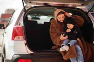 Young mother and child sitting in the trunk of a car and looking at mobile phone. Safety driving concept. photo