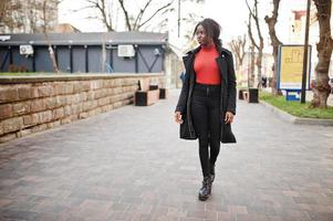 Portrait of a curly haired african woman wearing fashionable black coat and red turtleneck posing outdoor. photo