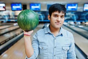 Stylish asian man in jeans shirt standing at bowling alley with ball at hand. photo