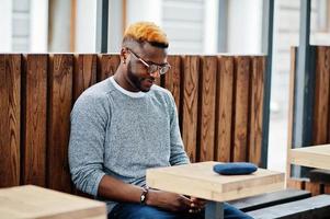 Stylish african american boy on gray sweater and glasses posed at street sitting on table wooden cafe. Fashionable black guy. photo