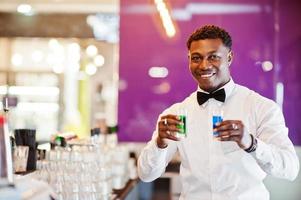 African american bartender at bar holding two coctail shots. Alcoholic beverage preparation. photo