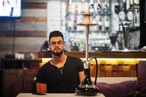 Stylish beard arabian man in glasses and black t-shirt smoking hookah indoor bar. Arab model having rest. photo