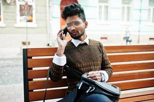 Portrait of young stylish indian man model pose in street, sitting on bench with handbag and smartphone. photo