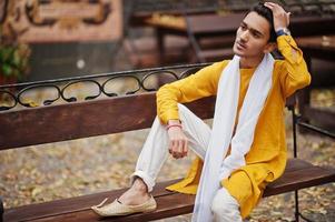 Indian stylish man in yellow traditional clothes with white scarf posed outdoor on bench. photo