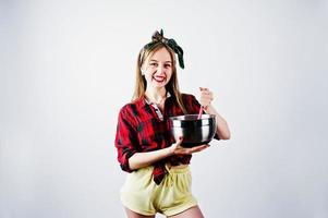 joven ama de casa divertida en camisa a cuadros y pantalones cortos amarillos pin up estilo con cacerola y cuchara de cocina aislado sobre fondo blanco. foto