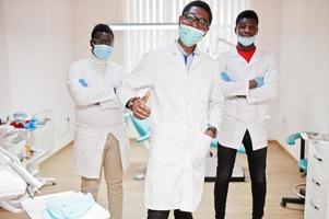 Three african american male doctor with crossed arms in dental clinic. Show thumb up. photo