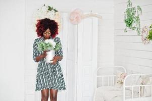 mujer modelo afroamericana alta de moda con pelo afro rojo en vestido posado en la habitación blanca con jarrón de flores en las manos. foto