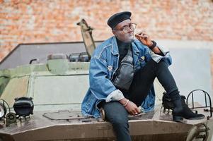 African american man in jeans jacket, beret and eyeglasses, smoking cigar and posed against btr military armored vehicle. photo