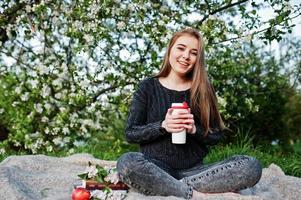 Young brunette girl at jeans sitting on plaid against spring blossom tree and holding tea thermos at hands. photo