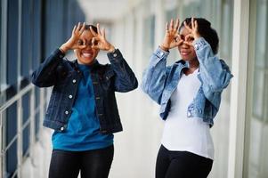 Two african woman friends in jeans jacket showing eyeglasses by fingers indoor together. photo