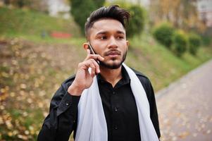 hombre con estilo indio vestido con ropa tradicional negra con pañuelo blanco posado al aire libre y hablando por teléfono móvil. foto