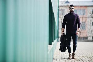 Fashion rich beard Arab man wear on turtle neck and sunglasses walking against green wall with jacket on hand. Stylish, succesful and fashionable arabian model guy. photo