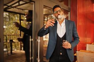 Handsome well-dressed arabian man smoke cigar with glass of whiskey at balcony of pub. photo
