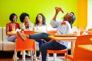 African woman with pizza sitting at restaurant against dark skinned girls. photo