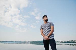 Handsome tall arabian beard man model at stripped shirt posed outdoor against lake and sky. Fashionable arab guy. photo