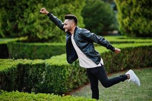 Stylish indian beard man at black leather jacket shows superman. India model posed at green garden of city. photo