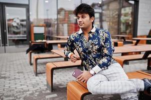 Attractive and handsome stylish indian man wear on shirt, holding mobile phone and sitting in the terrace of modern lounge cafe at his work break. photo