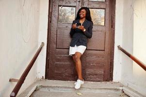 Success stylish african american woman in jacket and skirt against old wooden door with smartphone at hands. photo