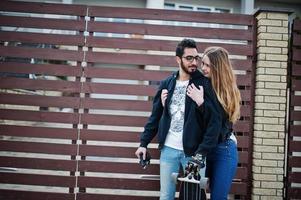 Cool multiracial couple hugs together with longboard against wooden fence. photo