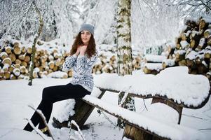 linda chica rizada con suéter y sombreros sentada en un banco en el parque forestal nevado en invierno. foto