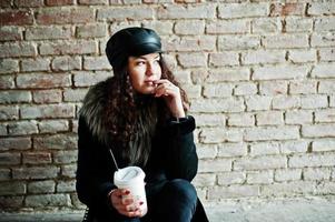 Curly mexican girl in leather cap and plastic cup of coffee at hand walking at streets of city. photo