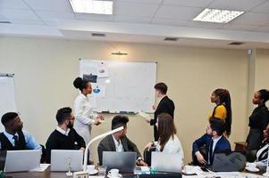 Mixed race business coach presenting report standing near whiteboard pointing on sales statistic shown on diagram and chart teach diverse company members gathered together in conference room. photo