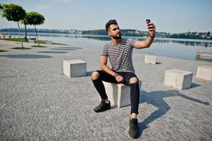 Handsome tall arabian beard man model at stripped shirt posed outdoor. Fashionable arab guy with mobile phone. photo