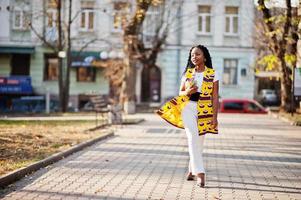 Stylish african american women in yellow jacket posed on street at sunny day with mobile phone at hand. photo